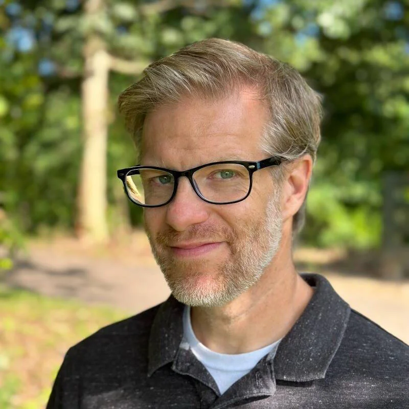 Headshot of white man with short blond hair, black glasses and a light beard in a black polo outside